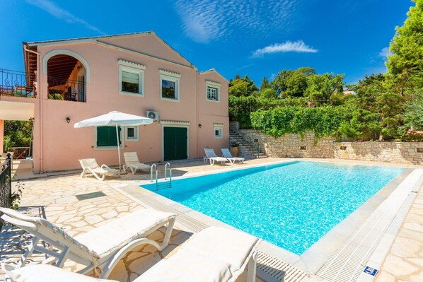 A two-story building with a swimming pool, outdoor seating, and a stone wall.