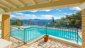 Ein Poolbereich mit freiem Blick auf das Meer und die Berge, umgeben von einer überdachten Terrasse mit Liegestühlen und Topfpflanzen.