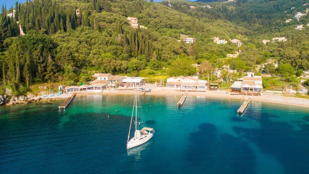 Ein Küstendorf mit kristallklarem türkisblauem Meer, einem am Steg vertäuten Segelboot und Gebäuden im mediterranen Stil.