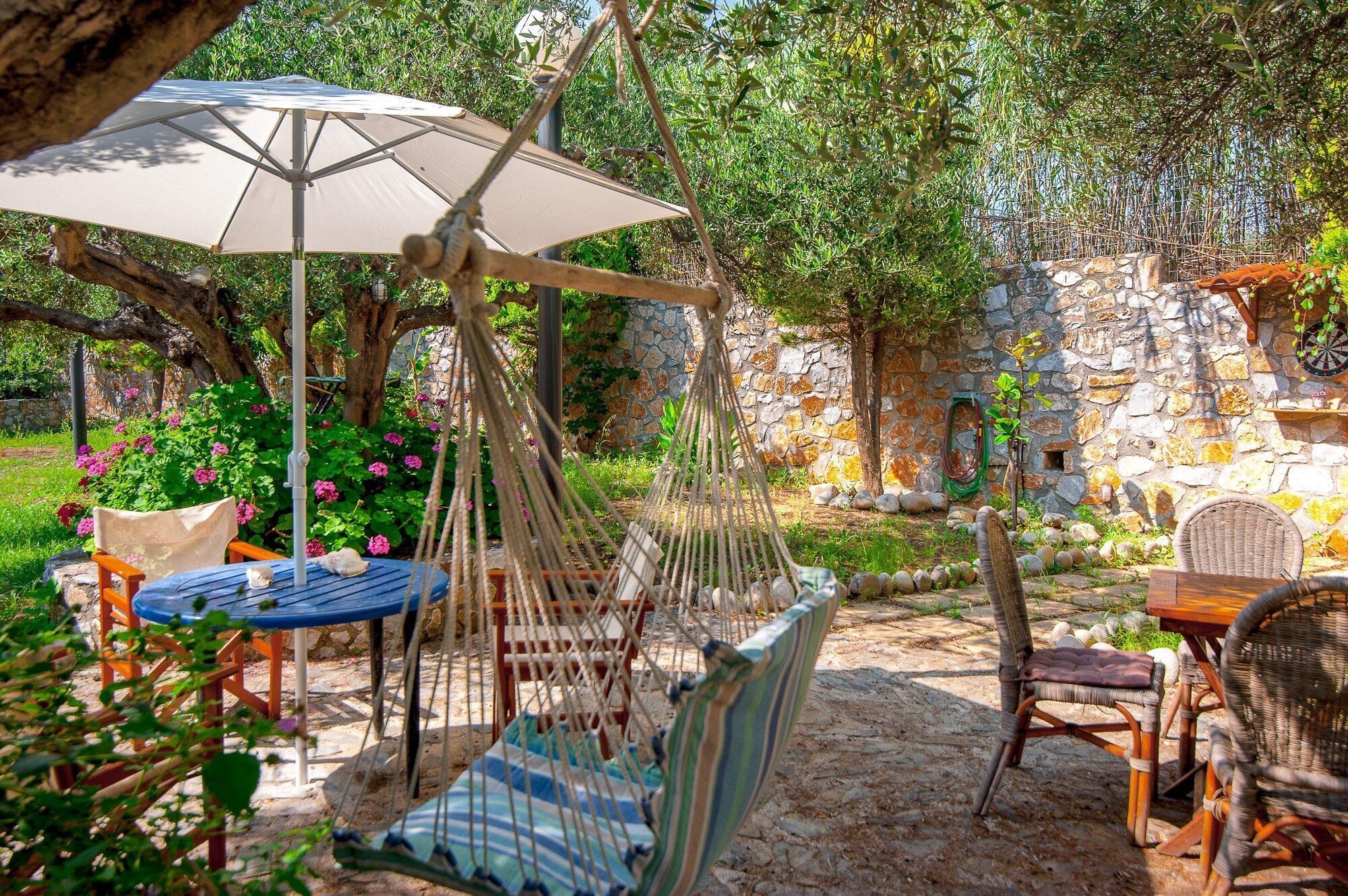 Un fauteuil suspendu, une table et une chaise placés sous un arbre dans un jardin agrémenté d’un mur de pierre et de fleurs.