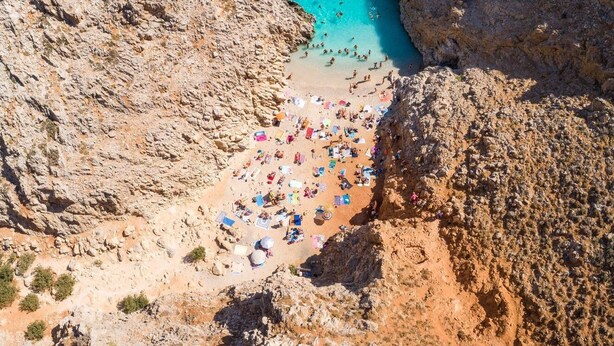 Um litoral rochoso com um mar turquesa límpido, uma praia de areia com guarda-sóis e algumas pessoas.