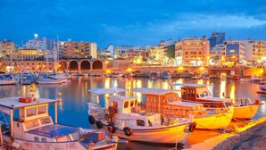 Uma marina com barcos atracados, um calçadão à beira-mar e edifícios ao fundo.