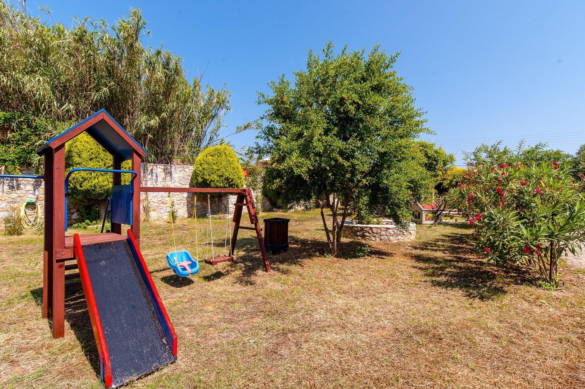 Un terrain de jeux avec un toboggan et des balançoires, entouré d’arbres et d’un mur de pierre.
