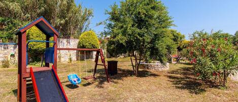Un terrain de jeux avec un toboggan et des balançoires, entouré d’arbres et d’un mur de pierre.