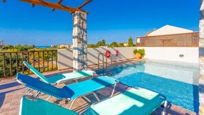 Uma piscina no terraço com cadeiras de descanso azuis, um pilar de pedra e vista para o mar.