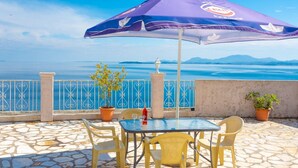 Une terrasse avec une table et des chaises, un parasol violet, des plantes en pot et une vue sur la mer et les montagnes.