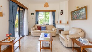 A living room with a beige sofa, a wooden coffee table, and a beige armchair. There is a window with blue curtains, a painting on the wall, and a clock. - Villa Eleni Large Private Pool Walk to Beach Sea Views A C Wifi Car Not Required Eco-friendly - 1840 (Pegeia)