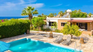 A swimming pool with lounge chairs, a building with a red roof, and palm trees. - Villa Eleni Large Private Pool Walk to Beach Sea Views A C Wifi Car Not Required Eco-friendly - 1840 (Pegeia)