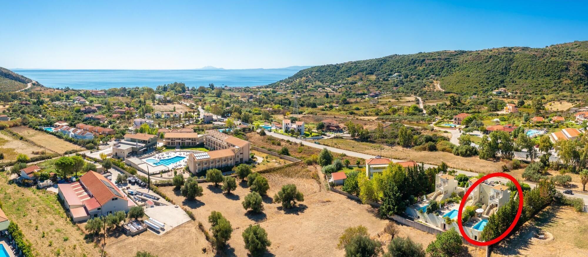 An aerial view of a coastal resort with multiple buildings, a large swimming pool, and a view of the sea.