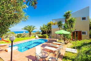 Piscina con sillas de descanso y una mesa, rodeada de vegetación y vista al mar.