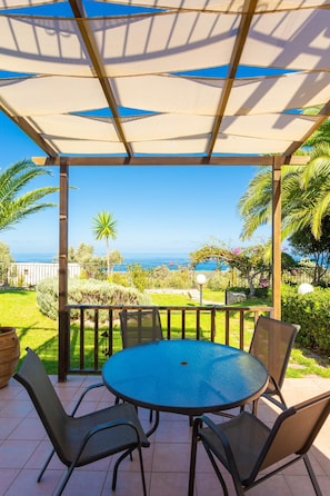 Un porche cubierto con una mesa redonda y sillas, con vistas a un jardín y al mar.