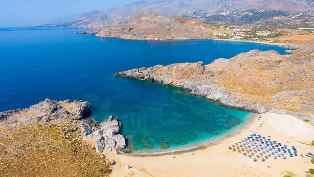 A coastal landscape with a rocky beach, clear blue waters, and distant mountains.