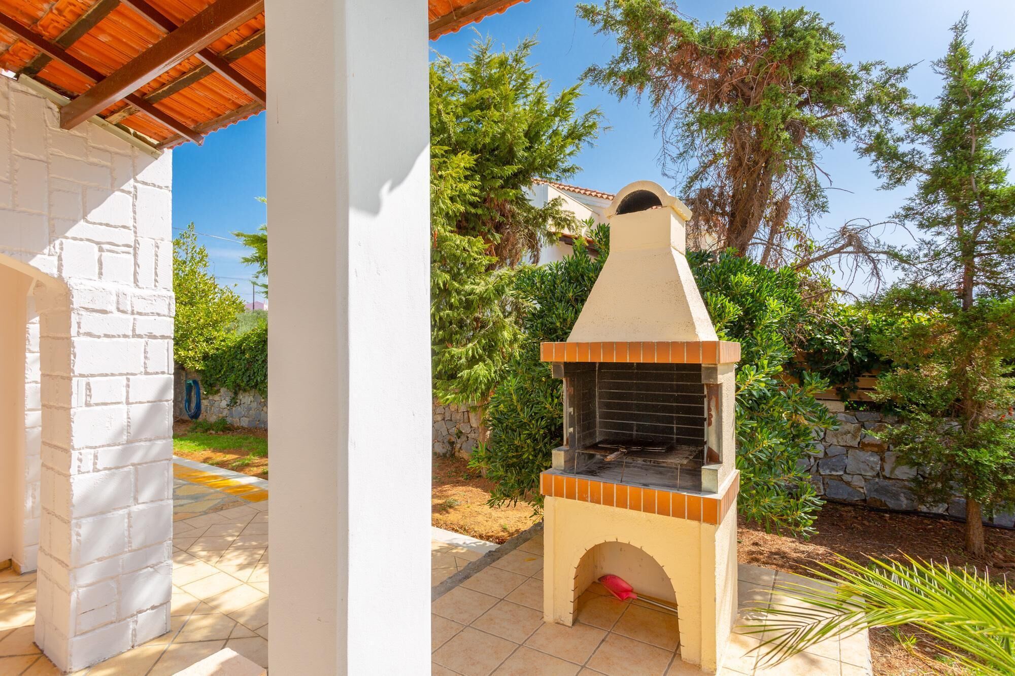 An outdoor brick barbecue grill with a chimney, set against a backdrop of trees and a clear sky.