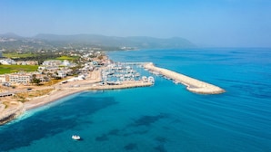 Vista aérea de una marina costera con barcos amarrados, un muelle que se adentra en el mar y una zona residencial con edificios y vegetación al fondo.