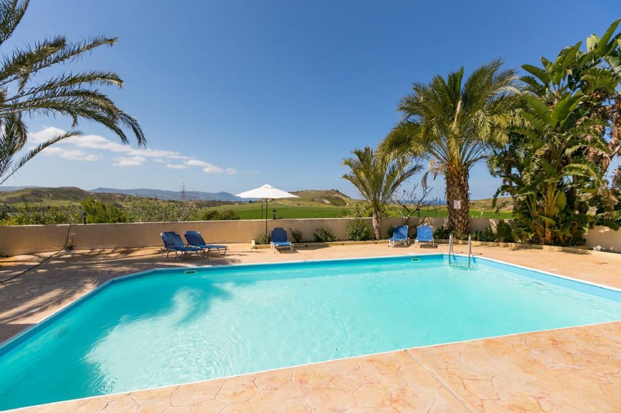 A swimming pool with clear blue water, surrounded by lounge chairs and palm trees, with a view of distant hills.