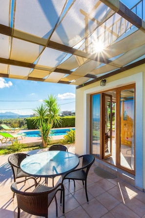 A covered outdoor area with a glass table and chairs, a pool, and a view of mountains in the background.