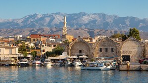Ein Hafen mit vor Anker liegenden Booten, eine Moschee mit einem Minarett und Berge im Hintergrund.