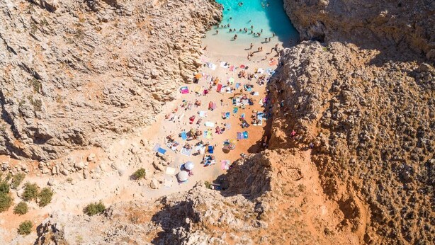 Eine felsige Küstenlinie mit einem Strandabschnitt, Sonnenschirmen und Menschen.