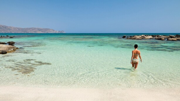 Een strand met helder ondiep water, een persoon in een bikini en rotsformaties in de verte.