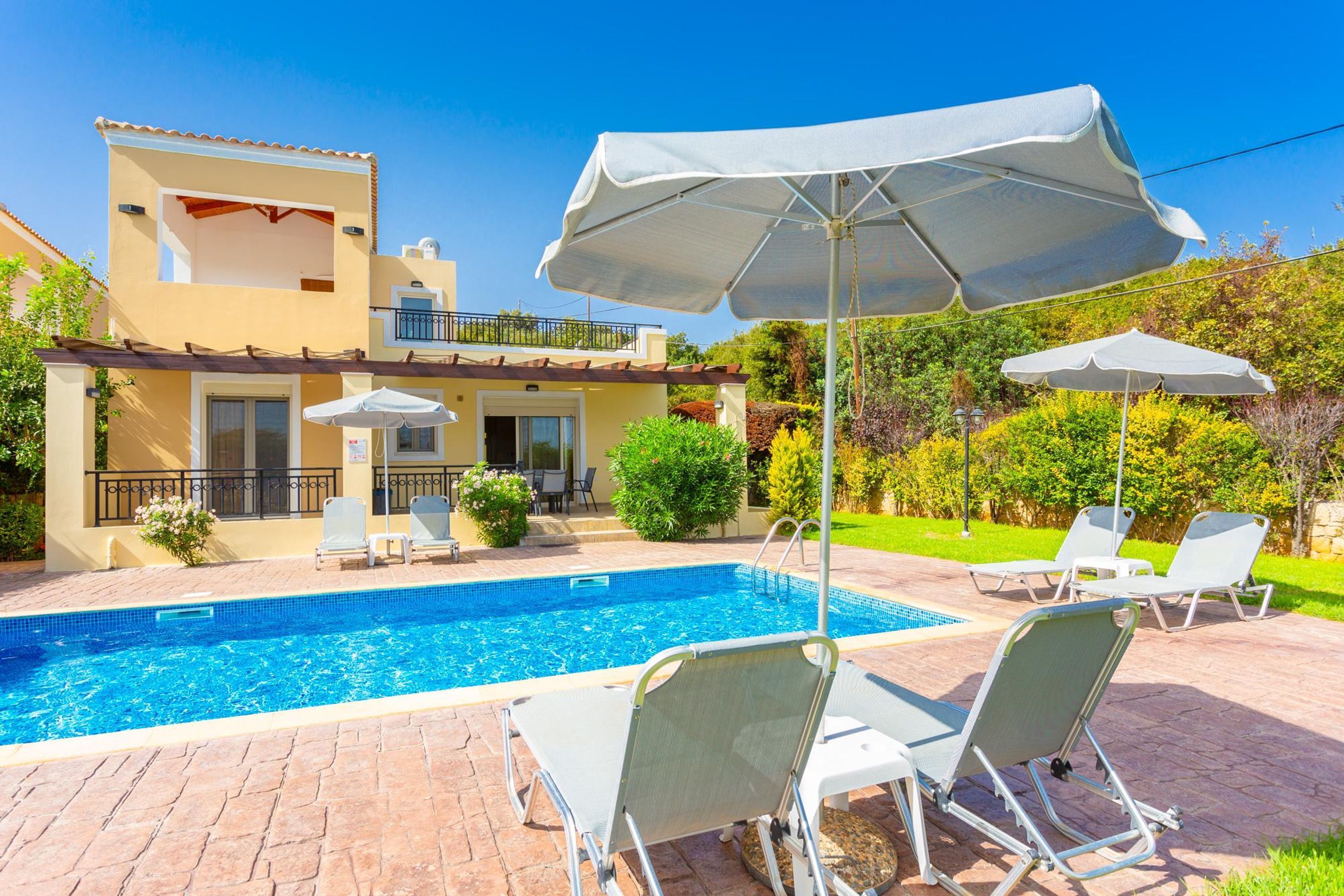 En swimmingpool med liggestole og parasoller, en bygning med balkon, og en velplejet have.