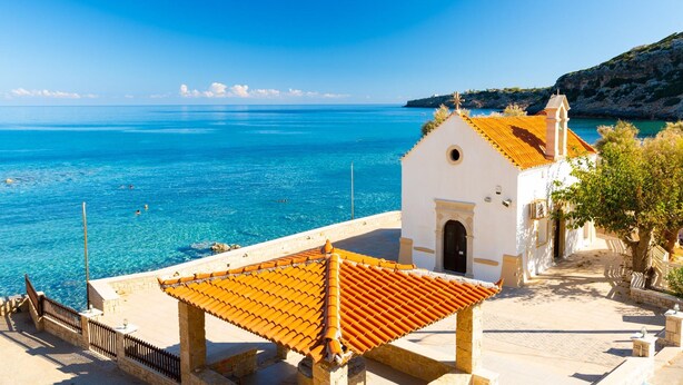 A coastal view with a white building featuring a red-tiled roof, a small church, and a clear blue sea.