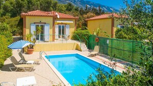 Ein Poolbereich mit zwei gelben Gebäuden, einem blau-wei ß gestreiften Sonnenschirm und einem Berg im Hintergrund.