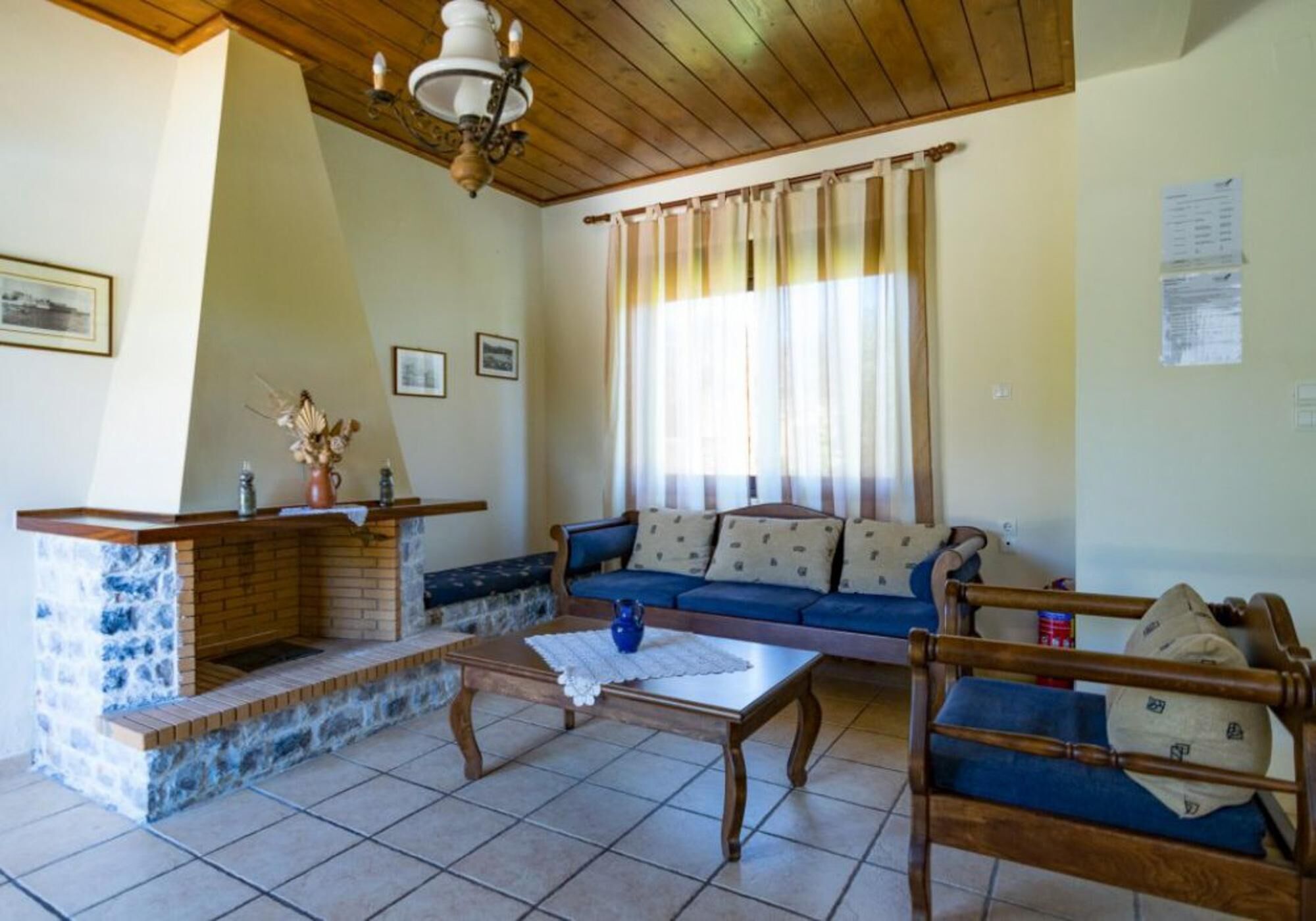 Un salón con chimenea de ladrillo, un sofá azul, una mesa de centro de madera y una ventana con cortinas.