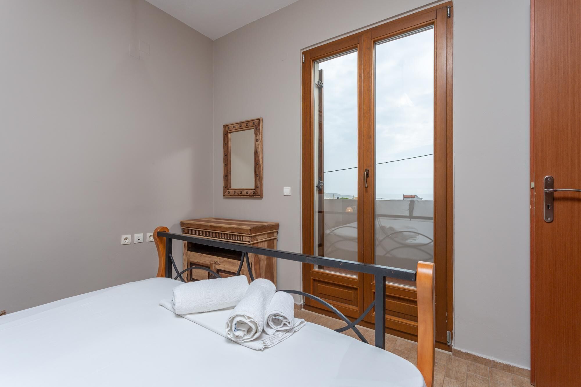 A bedroom with a bed, a wooden chest, a mirror, and a sliding glass door leading to a balcony.