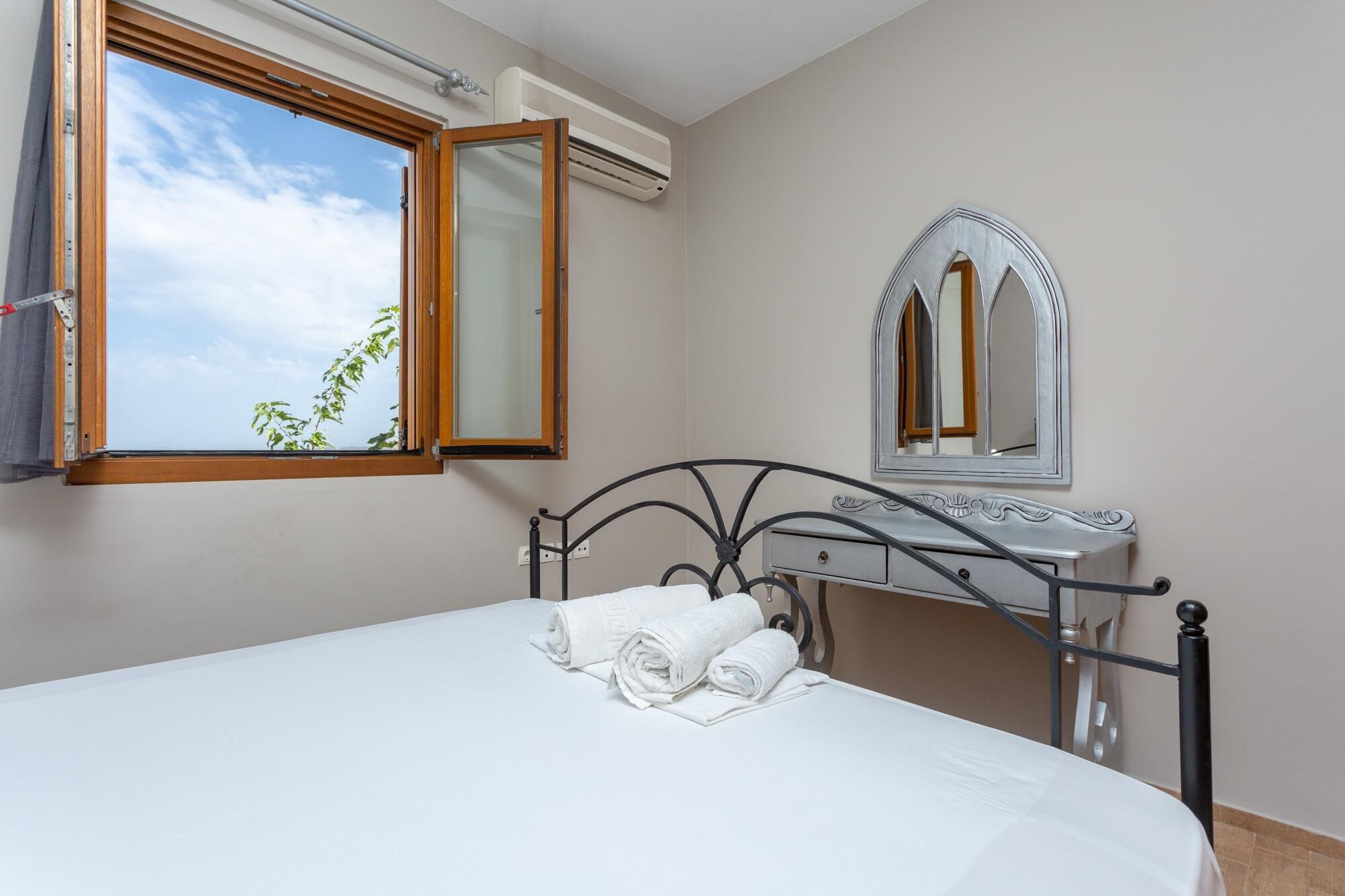 Een slaapkamer met een bed, een raam met een open gordijn, een airconditioningapparaat, een kast en een spiegel.