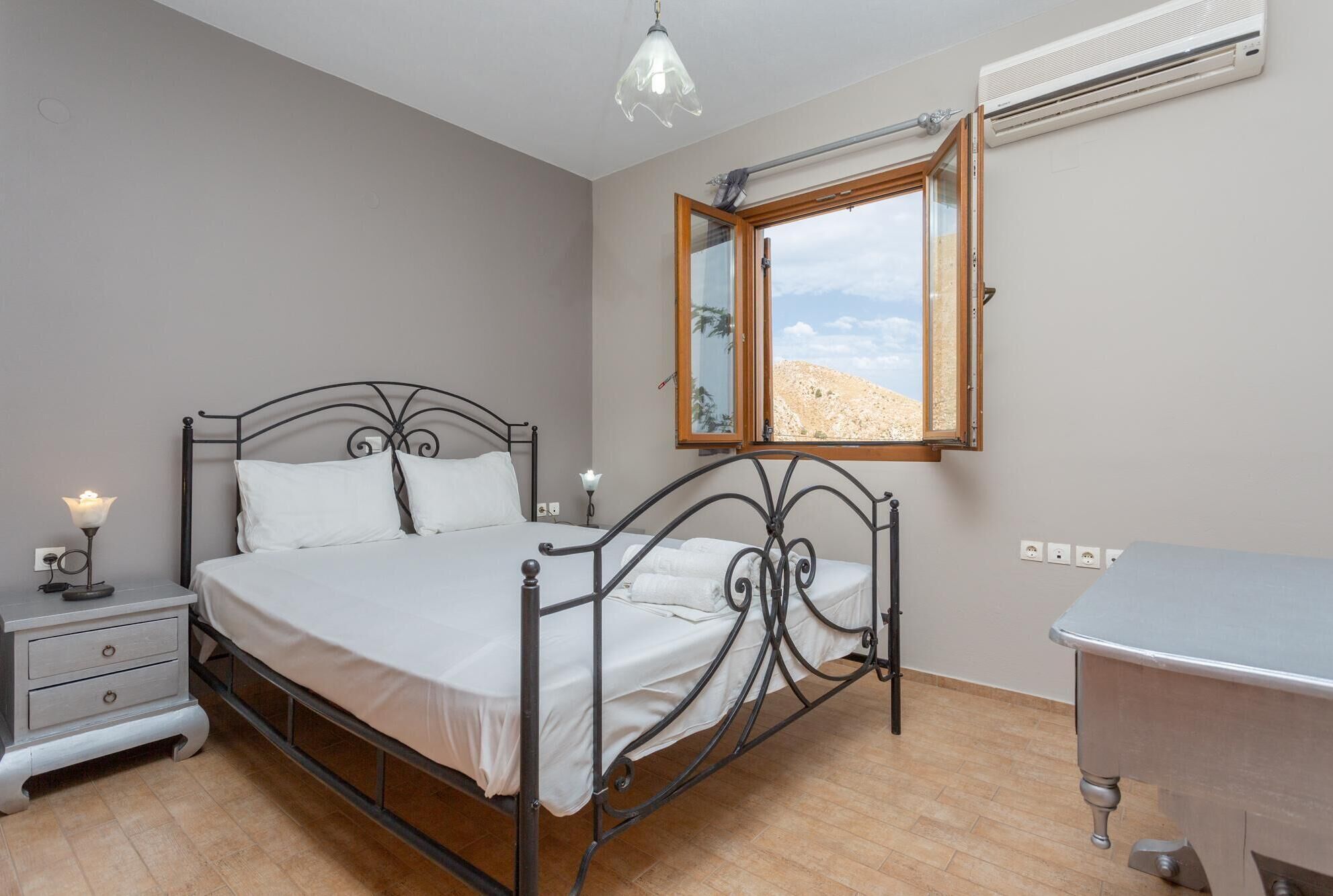 Un dormitorio con cama, mesita de noche, lámpara y ventana con vistas a una colina.