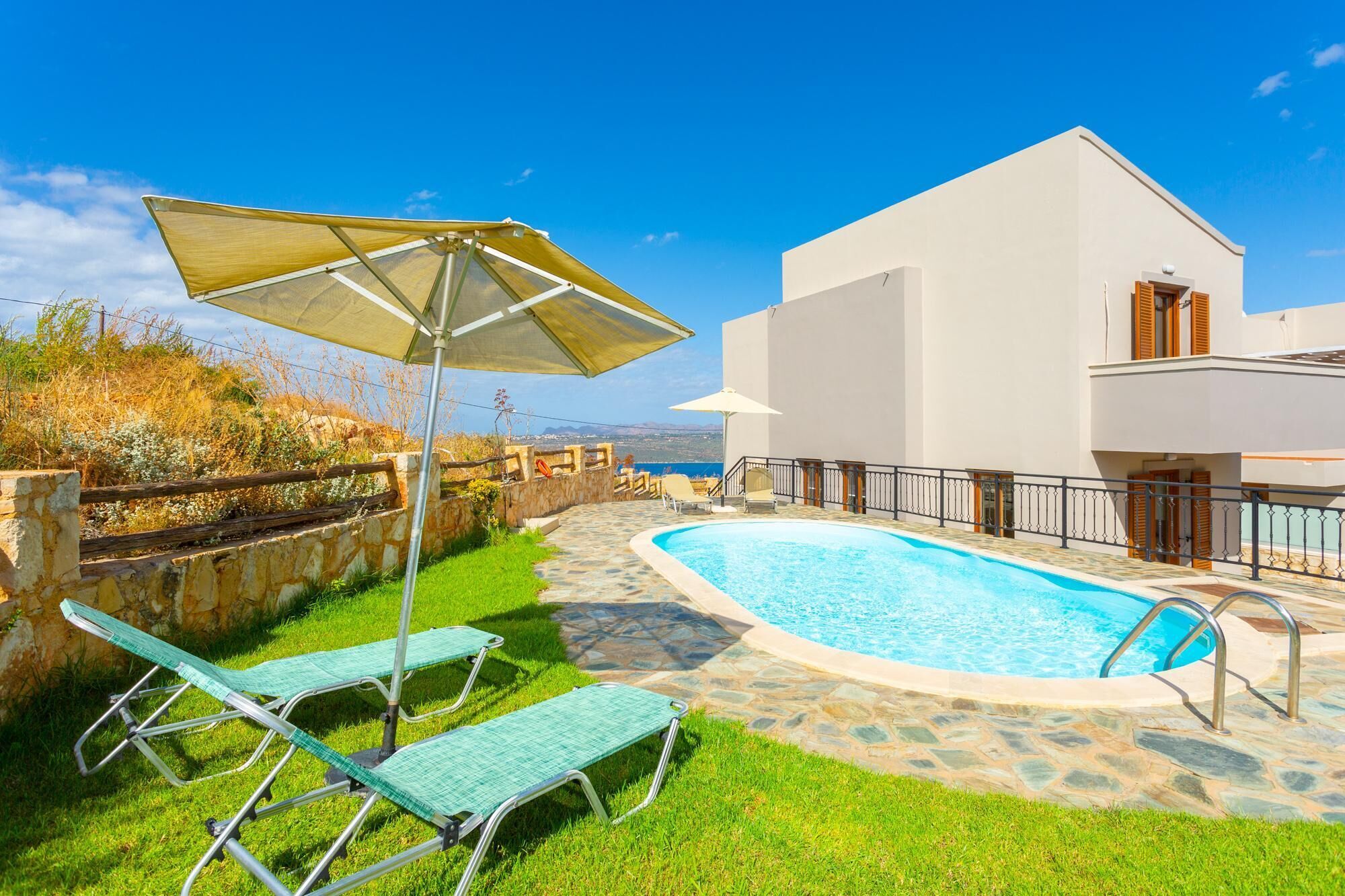 A swimming pool with lounge chairs and an umbrella, surrounded by a stone wall and a white building with a balcony.