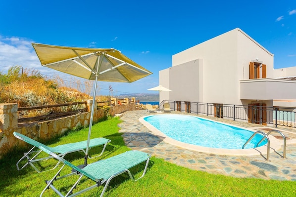 A swimming pool with lounge chairs and an umbrella, surrounded by a stone wall and a white building with a balcony.
