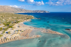 An aerial view of a coastal area with a beach, clear blue waters, and mountainous terrain in the background.