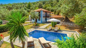 Une maison à deux étages avec une piscine, entourée d’une végétation luxuriante et d’oliviers.