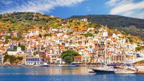 Une ville côtière aux bâtiments blancs et aux toits rouges, un port où sont amarrés des bateaux, et une colline verdoyante.