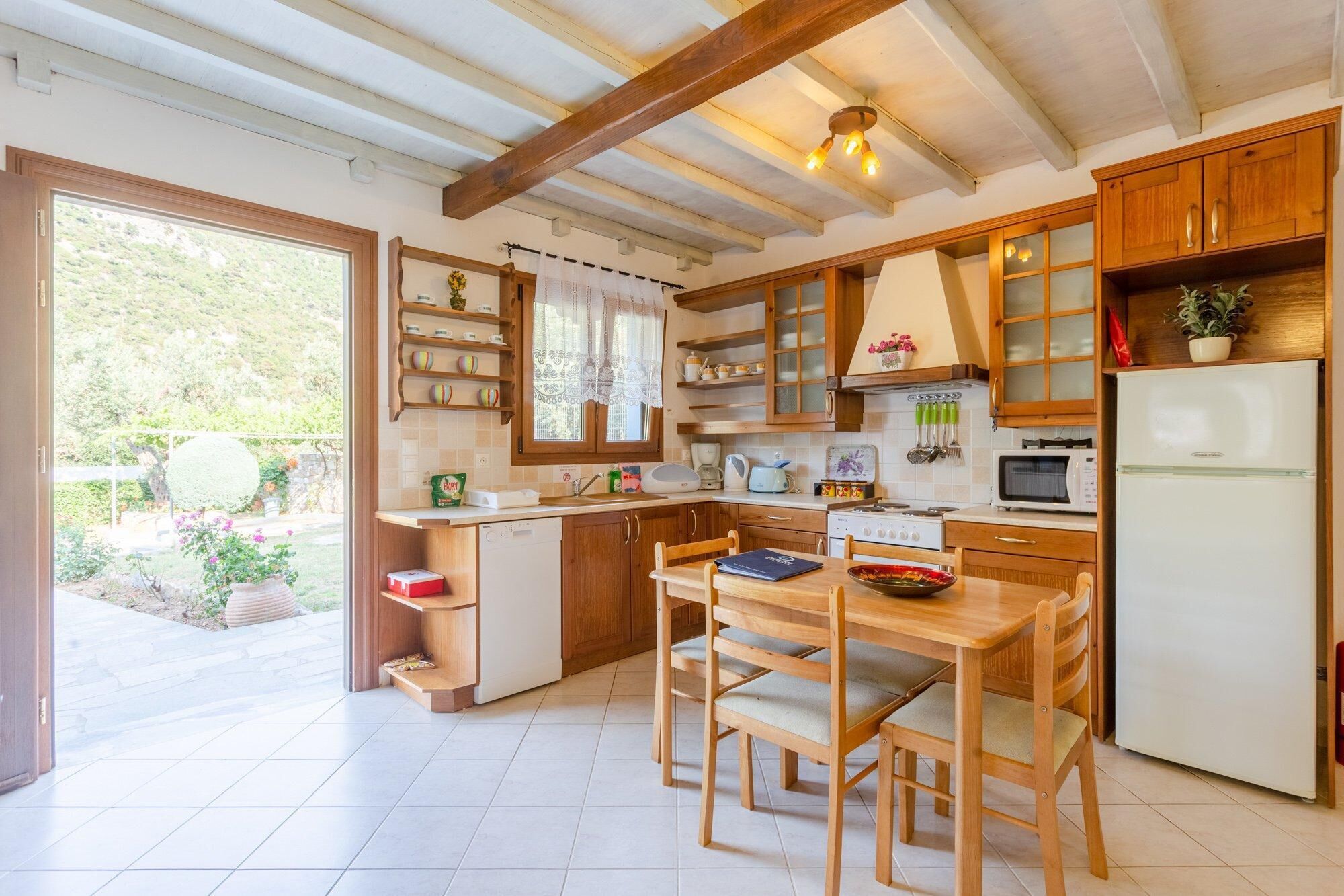 Une cuisine avec des meubles en bois, une table à manger avec des chaises, un réfrigérateur et une fenêtre donnant sur un jardin.