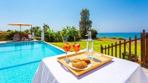 A poolside table with a tray of food and drinks, a bottle of water, and a view of the ocean. - Kalisto Beach Villa Large Private Pool Walk to Beach Sea Views A C Wifi Car Not Required - 2137 (Ayia Marina Chrysochous)