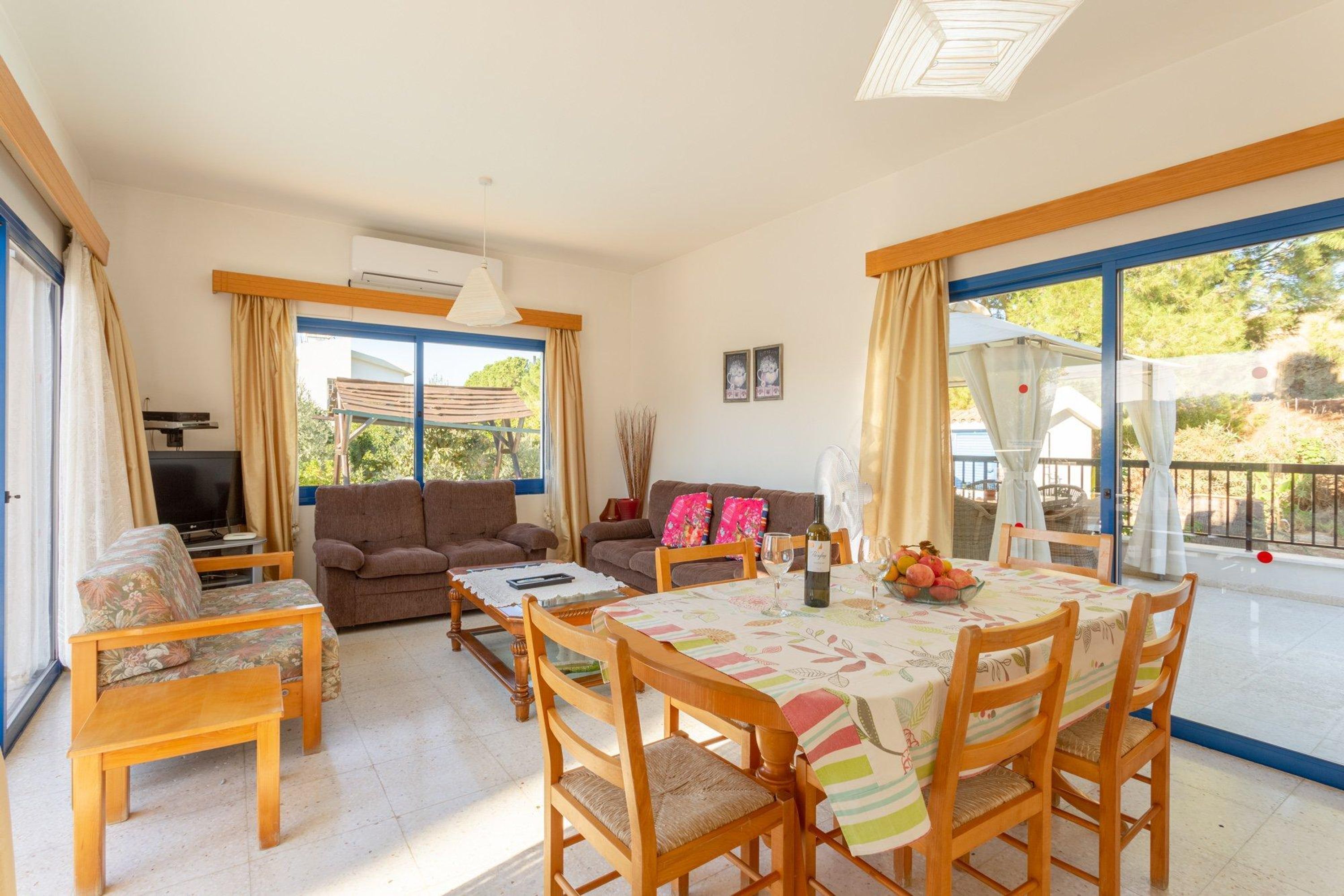 A spacious living area with a sofa, a dining table with chairs, a television, and large windows with curtains.