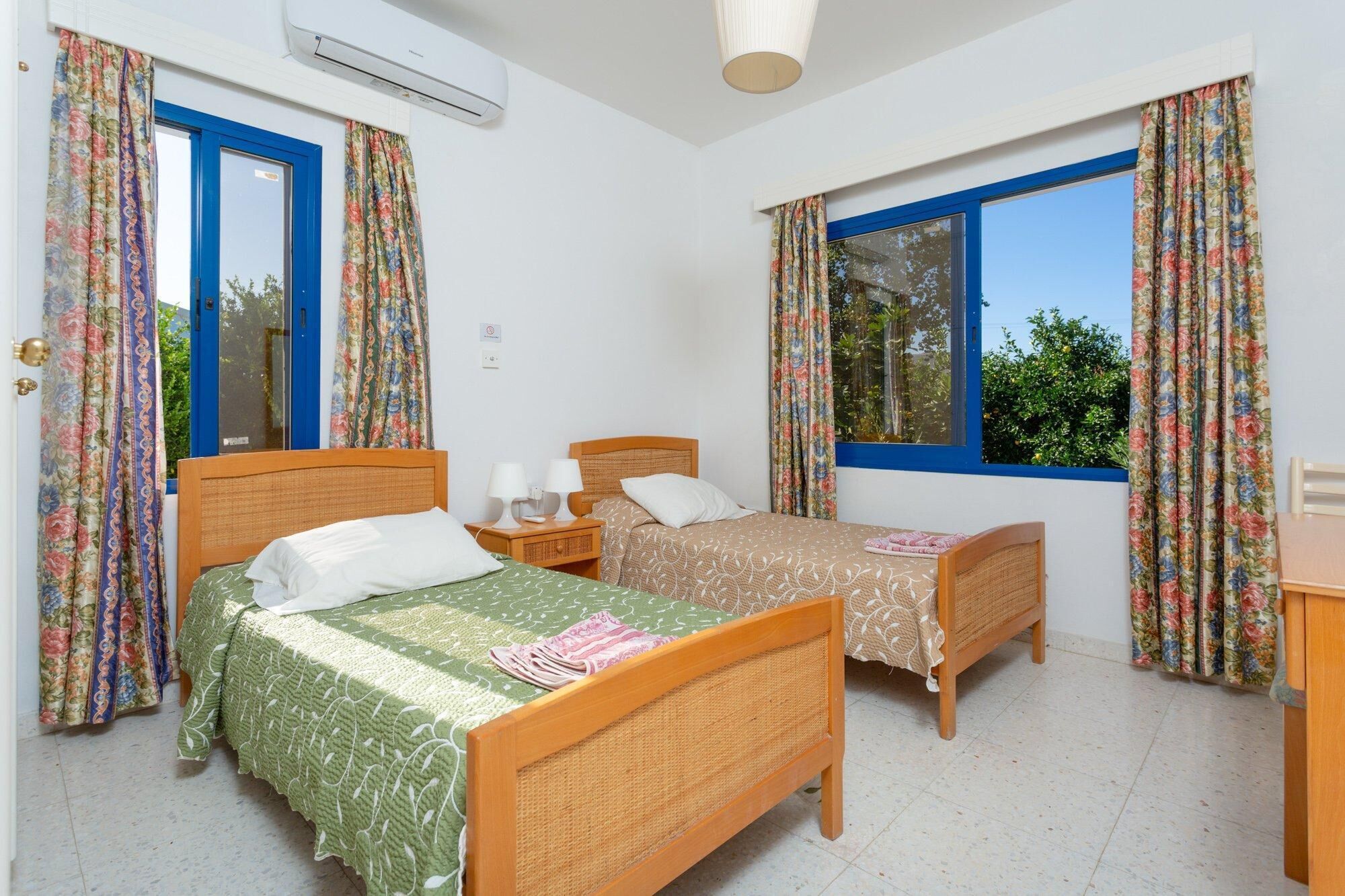 A bedroom with two single beds, a wooden nightstand with lamps, a small table, and a window with curtains.