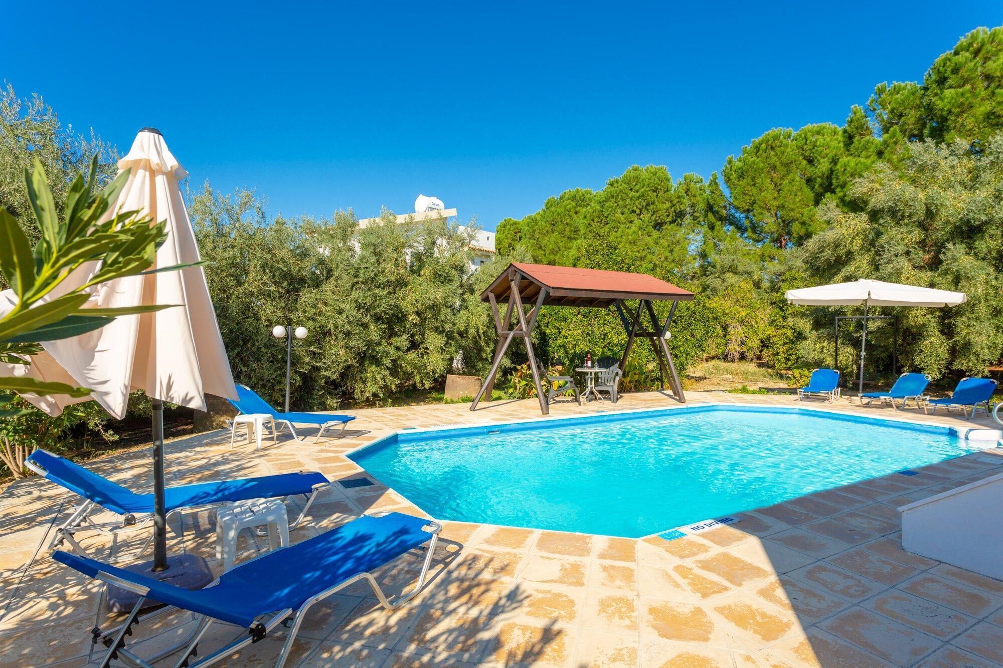 A swimming pool with lounge chairs and umbrellas, surrounded by greenery and a clear sky.