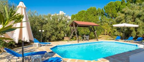 A swimming pool with lounge chairs and umbrellas, surrounded by greenery and a clear sky.