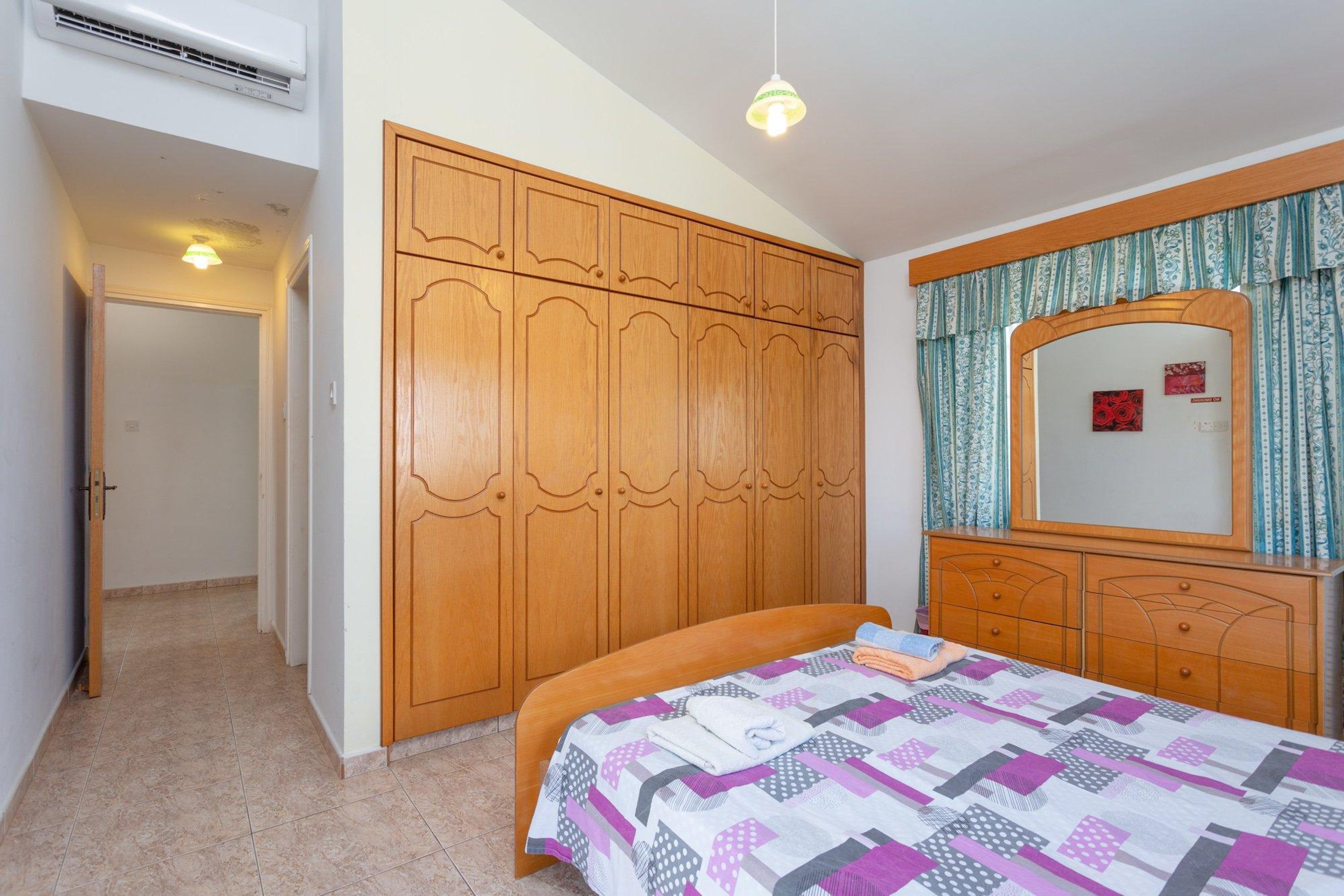 A bedroom with a wooden wardrobe, a bed with a patterned bedspread, a dresser with a mirror, and a hanging light fixture.