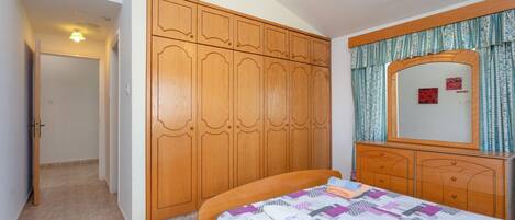 A bedroom with a wooden wardrobe, a bed with a patterned bedspread, a dresser with a mirror, and a hanging light fixture.