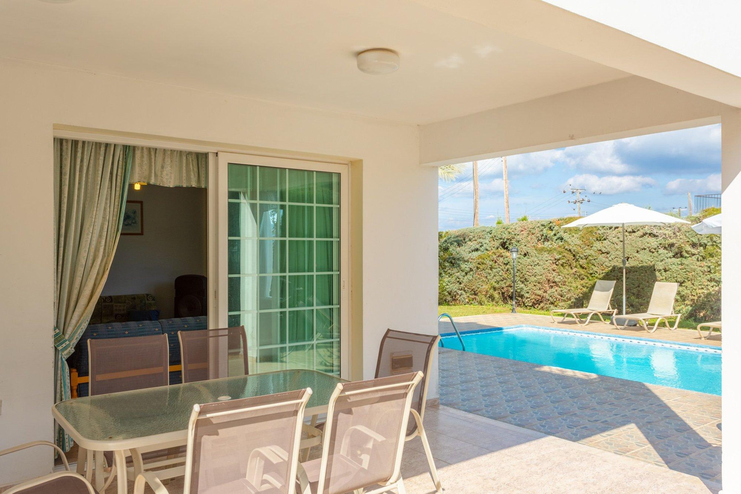An outdoor pool area with a glass table and chairs, a sliding glass door, and a view of the pool and greenery.