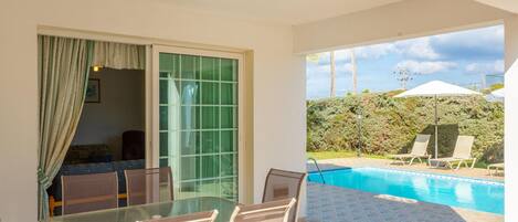 An outdoor pool area with a glass table and chairs, a sliding glass door, and a view of the pool and greenery.