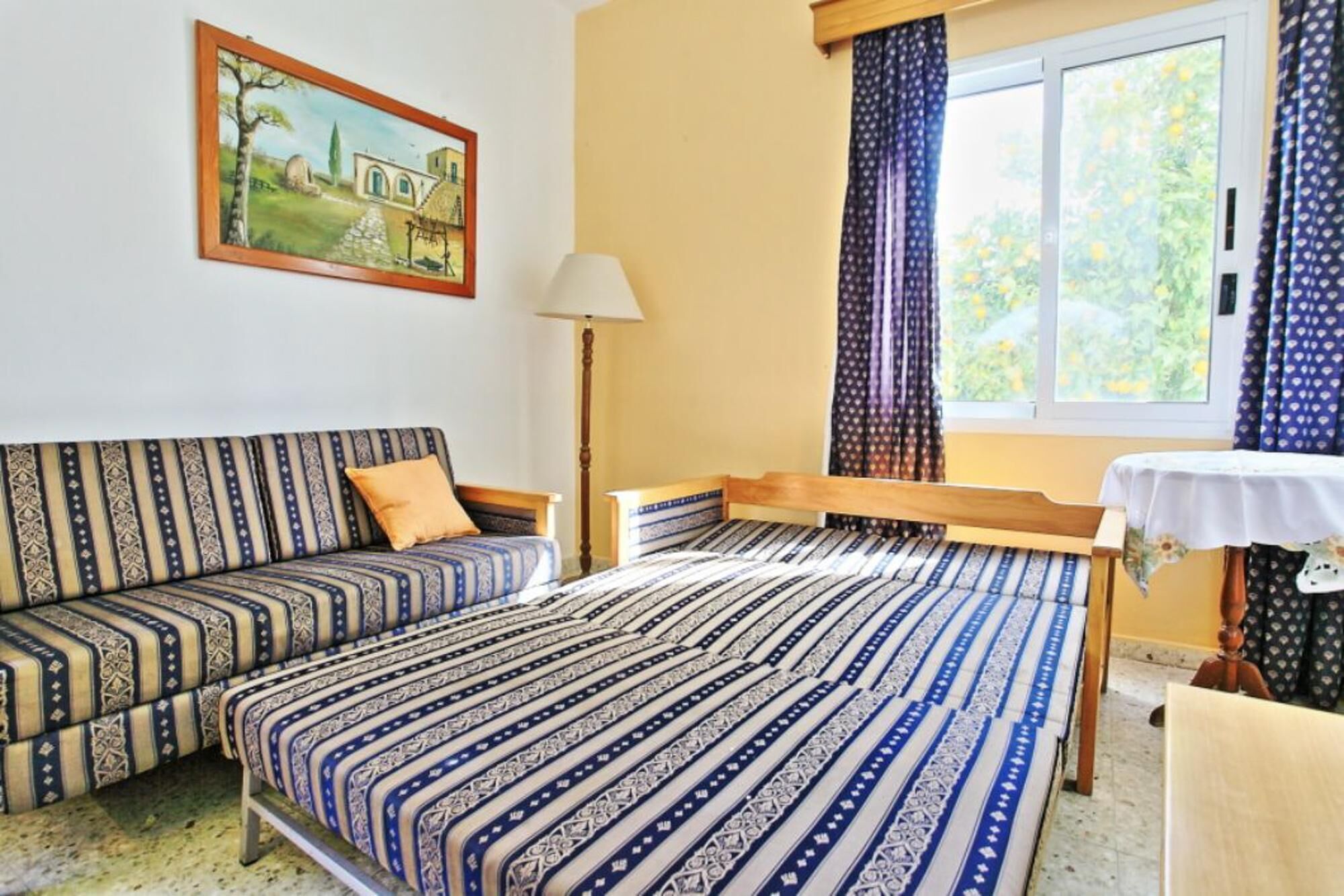 A room with a sofa and a bed, a painting on the wall, a window with curtains, and a table with a tablecloth.
