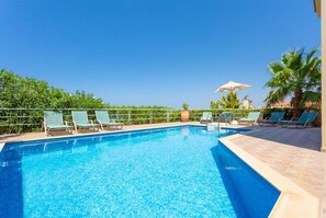 A clear blue pool with lounge chairs and umbrellas, surrounded by greenery and a clear sky.