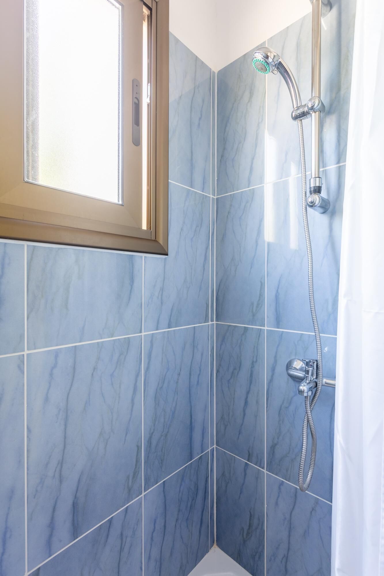 Un baño con baldosas de mármol azul, una ducha con cabezal de ducha manual y un ventanal con cristal satinado.