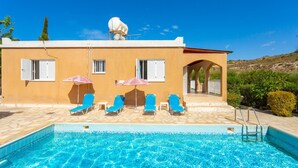 Uma área à beira da piscina, com uma piscina azul cristalina, um edifício com fachada bege e uma área de estar coberta com espreguiçadeiras azuis e guarda-sóis listrados em vermelho e branco.