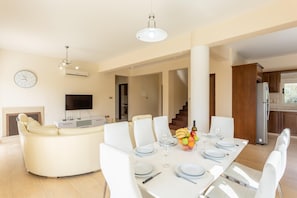 A modern dining area with a white table set for six, a sofa, and a TV in the background.